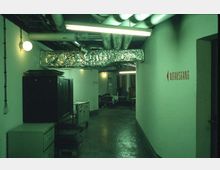 Ein unterirdischer Flur mit weiß gestrichenen Wänden, sichtbarer Rohr- und Lüftungstechnik an der Decke sowie Neonbeleuchtung. An der rechten Wand befindet sich ein roter Schriftzug mit der Aufschrift „Notausgang“ und einem Pfeil. Auf dem Boden stehen Metallmöbel und Rollwagen.