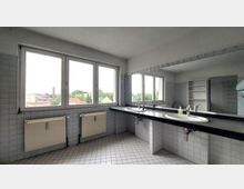 Großes Badezimmer mit weiß gefliesten Wänden und Boden, einem langen Waschtisch mit zwei Waschbecken und einem großen Spiegel darüber. Drei Fenster bieten Blick auf eine städtische Umgebung mit Häusern, Bäumen und einem Schornstein.