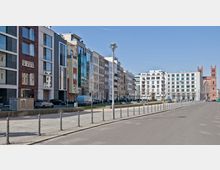 Straße mit modernen Wohngebäuden auf der linken Seite, die durch unterschiedliche Fassadenmaterialien und Farben geprägt sind, vor einem Gehweg mit Pollern. Im Hintergrund steht eine rote Backsteinkirche mit zwei Türmen, flankiert von weiteren weißen Gebäuden.