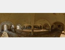 Ein heruntergekommener, gewölbter Innenraum eines historischen Gebäudes mit mehreren Rundbögen, bröckelnden Ziegelwänden und Stützen aus Stein. Tageslicht fällt durch eine offene Tür und Fensteröffnungen, während der Boden mit Schmutz und Baumaterialien bedeckt ist.