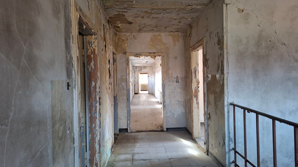 Langer, leerer Flur in einem verfallenen Gebäude mit abblätternder Farbe und rissigem Putz an Wänden und Decke, mehreren offenen Türrahmen und Fliesenboden. Am rechten Rand ist ein Treppengeländer zu sehen, am Ende des Flurs fällt Tageslicht durch ein Fenster.