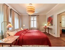 Gemütlich eingerichtetes Schlafzimmer mit einem Doppelbett, das mit einer roten Decke bedeckt ist, zwei Nachttischen mit dekorativen Lampen und einem großen Fenster mit cremefarbenen Vorhängen. An der Wand hängen ein farbenfrohes Blumenbild und ein großer, runder Deckenlampenschirm, der warmes Licht spendet.