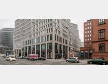 Straßenszene mit modernen Bürogebäuden, darunter ein großes Eckgebäude mit horizontal gestreiften Fassaden und großen Glasfenstern im Erdgeschoss. Im Hintergrund ein weiteres rundes Bürogebäude und ein rotes Backsteingebäude mit großen Fenstern; parkende Autos entlang der Straße.