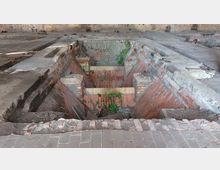 Innenraum einer verlassenen Industriehalle mit sichtbarer, rechteckiger Vertiefung aus roten Ziegelsteinen, umgeben von Betonflächen. Die Vertiefung weist Spuren von Verfall auf, mit eingewachsenem Grün und bröckelnden Ziegelwänden.