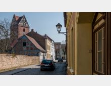 Eine gepflasterte, historische Straße mit Fachwerk- und Backsteinhäusern, gesäumt von einer Ziegelmauer links und einem gelb verputzten Gebäude rechts. Im Hintergrund ist ein Kirchturm aus rotem Backstein sichtbar, während ein geparktes Auto die Straße belebt.