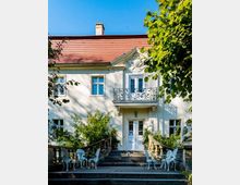Historisches Gebäude mit heller Fassade und rotem Ziegeldach, umgeben von Bäumen. Eine symmetrische Treppe mit Geländern führt zu einem Eingang mit dekorativem Balkongeländer, davor stehen weiße Tische und Stühle.