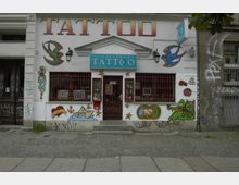 Fassade eines Tattoo-Studios mit dem Schriftzug "Tattoo" und einem Schild mit der Aufschrift "Loxodrom Tattoo Piercing". Die weiße Wand ist mit bunten Wandmalereien wie einem Segelschiff, Schwalben, Rosen und weiteren Motiven gestaltet. Links und rechts befinden sich graffitibeschmierte Wände.