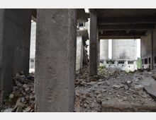 Innenansicht eines verlassenen, teils zerstörten Industriegebäudes mit Betonpfeilern und Schutt auf dem Boden. Im Hintergrund sind große, graue Silos und beschädigte Wände zu sehen.