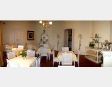 Ein kleiner, eleganter Speiseraum mit hellen Wänden, Holzboden und einer Deckenleuchte. Es stehen weiße Tische mit gelben Tischdecken und geflochtenen Stühlen, umgeben von Regalen mit dekorativen Gegenständen wie Silbergeschirr und Blumendekorationen.