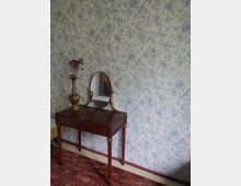 Ein kleiner, antiker Holztisch mit einer ovalen Spiegelhalterung steht vor einer blassblauen Tapete mit floralem Muster. Auf dem Tisch steht eine verzierte Vase mit roten Blüten, und der Boden ist mit einem orientalischen Teppich bedeckt.