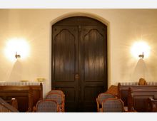 Holztür mit dunkler Lackierung in einem Kirchenraum, flankiert von schlichten, leuchtenden Wandleuchten. Im Vordergrund befinden sich hölzerne Kirchenbänke und gepolsterte Stühle, die zur Einrichtung beitragen.