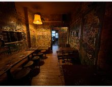 Schmaler, schwach beleuchteter Innenraum einer Bar oder Kneipe mit Holzboden, einfachen Tischen und Hockern sowie einer hängenden Lampe. Die Wände und Türen sind dicht mit Graffiti, Stickern und Kritzeleien bedeckt, im Hintergrund führt ein Durchgang in einen weiteren Raum.