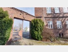 Ein historisches Backsteingebäude mit weißen Fenstern und dekorativen Metallgittern; links ein großes, offenes Tor mit einem Metallgitter und Ziegelrahmen. Hohe grüne Büsche und gepflasterter Boden flankieren den Eingang, dahinter ist eine asphaltierte Fläche sichtbar.