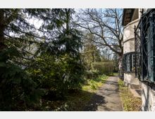 Seitenansicht eines Wohnhauses mit verzierten Metallgittern an den Fenstern, umgeben von Bäumen und Sträuchern. Ein schmaler gepflasterter Weg führt durch einen Garten mit Blumen und dichter Vegetation.