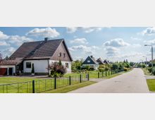 Eine ruhige Wohnstraße mit freistehenden Einfamilienhäusern, gepflegten Gärten und Gehwegen, die von Metallzäunen abgegrenzt sind. Der Himmel ist klar mit einigen Wolken, und die Straße ist mit gepflasterten Steinen ausgelegt.