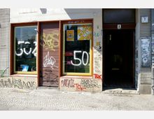 Fassade eines Gebäudes mit zwei großen Schaufenstern, von denen eines mit Graffiti besprüht ist, sowie einer offenen Eingangstür rechts daneben. Die Wände und Fensterrahmen sind mit weiteren Graffiti bedeckt, und der Boden besteht aus einer gepflasterten Gehsteigfläche.