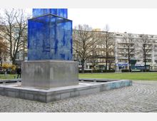 Skulptur auf einem öffentlichen Platz mit einem hohen, transparent-blauen Glaselement auf einem metallischen Sockel. Im Hintergrund sind Wohngebäude, Bäume und eine Grünfläche zu sehen.
