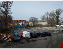 Eine leere gepflasterte Fläche mit geparkten Autos im Vordergrund, umgeben von Bäumen und großen Gebäuden im Hintergrund, darunter ein historisches Backsteingebäude und moderne Hochhäuser. Graffiti-bedeckte Mauern und Bauzäune grenzen das Gelände ab; ein Autovermietungswerbeschild ist links sichtbar.