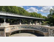Gewölbte Steinbank mit Graffiti vor einer erhöhten Metallbrücke, darunter ein Fluss und üppige grüne Bäume im Hintergrund bei sonnigem Wetter.