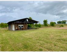 Eine einfache Holzhütte mit einem Flachdach und einer überdachten Veranda steht auf einer weiten Wiese. Im Hintergrund befinden sich Büsche, Bäume und ein bewölkter Himmel.