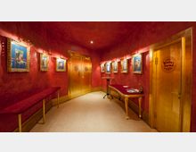 Roter Innenflur mit goldenen Türen und einer warmen Beleuchtung, geschmückt mit gerahmten Bildern an den Wänden. Ein schmaler roter Konsolentisch und ein runder Stehtisch stehen am Rand, während auf einer goldenen Tür das Logo des "Wintergarten Varieté" sichtbar ist.