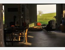 Wohnraum in einem rustikalen Gebäude mit Sichtbeton und unverputzten Wänden, ausgestattet mit einem Holztisch, Stühlen, einer schwarzen Ledersesselgruppe und einem Kamin. Ein großes Fenster bietet Blick auf eine grüne Wiese mit einem Feld und einem Kürbis auf der Fensterbank.