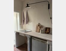 Kleine Küchenzeile mit rustikaler Gestaltung, bestehend aus einem weißen Spülbecken, einer Holzarbeitsplatte und einem kleinen Kühlschrank darunter. An der grob verputzten Wand hängen zwei gestreifte Geschirrtücher, und darüber ist eine schwarze, schwenkbare Wandlampe montiert; daneben stehen Utensilien und ein Holzbrett.