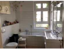 Helles Badezimmer mit einer Badewanne, einem Waschbecken und einer Toilette. Die Wände sind teils gefliest und teils mit weißen Blumenmustern verziert; ein großes Fenster lässt Tageslicht herein.