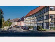 Wohnstraße mit mehrstöckigen Wohnhäusern im traditionellen Baustil und roten Ziegeldächern. Am Straßenrand parken Autos, und ein Schild weist auf eine Tempo-30-Zone hin; Gehwege sind von Bäumen und Hecken gesäumt.