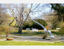 Ein Park mit einer großen, blattlosen Birke im Zentrum, umgeben von Blumenbeeten und Büschen. Im Vordergrund steht eine geschwungene Metallskulptur, dahinter sind weiße Bänke und dichtes Gebüsch zu sehen.