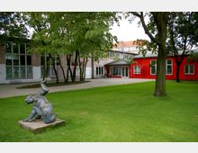 Außenbereich mit einer Grünfläche, einigen Bäumen und einer modernen, farbenfrohen Gebäudefassade, teils in Rot gestrichen. Im Vordergrund steht eine Bronzeskulptur auf einem Sockel, die eine menschliche Figur darstellt.