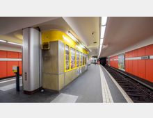 Eine U-Bahn-Station mit orangefarbenen Wandpaneelen, einem gelben Kiosk mit beleuchteten Auslagen in der Mitte des Bahnsteigs und einem Gleis auf der rechten Seite. Im Hintergrund ist ein Schild mit der Aufschrift „Rudow“ sichtbar.