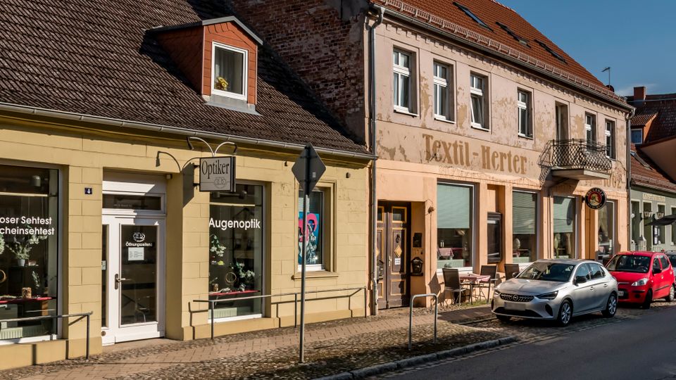 Straßenzug in einer Kleinstadt mit niedrigen, aneinandergebauten Wohn- und Geschäftshäusern mit Ziegeldächern und Ladenfronten, darunter ein Optiker und ein älteres Geschäft mit verblasster Fassadenaufschrift. Entlang der Straße stehen mehrere geparkte Autos, davor verläuft ein schmaler Gehweg mit Kopfsteinpflaster und Geländern.