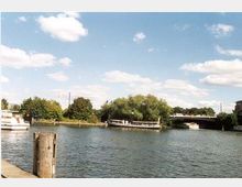 Flusslandschaft mit einer kleinen Insel, auf der dichte Bäume stehen, und einer Brücke im Hintergrund. Im Vordergrund sind Wasserflächen, ein Holzpfosten und ein Boot an einem Anlegesteg sichtbar.