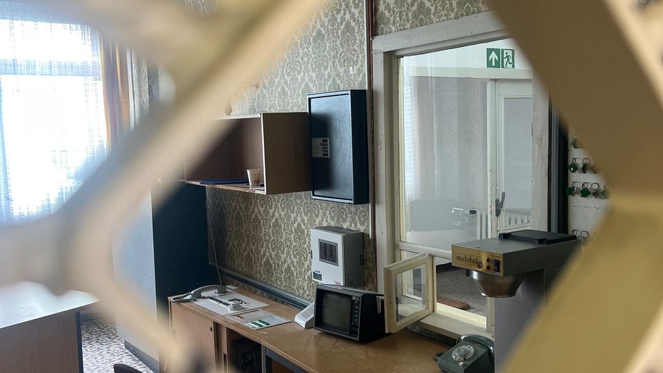 Blick durch ein Gitter in einen kleinen, altmodischen Büroraum mit gemusterter Tapete und Fliesenboden. Entlang der Wand stehen Schreibtische mit Telefon und technischen Geräten; daneben eine Glastür mit Fluchtwegzeichen und ein großes Fenster mit Vorhang.