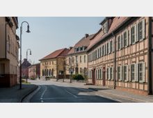 Straßenszene in einer Altstadt mit Fachwerkhäusern auf der rechten Seite und einem gelben Gebäude mit Arkaden auf der linken Seite, das als Veranstaltungsgebäude dient. Die gepflasterte Straße ist von Laternen und kleinen Bäumen gesäumt, während die Umgebung einen ruhigen, städtischen Charakter zeigt.