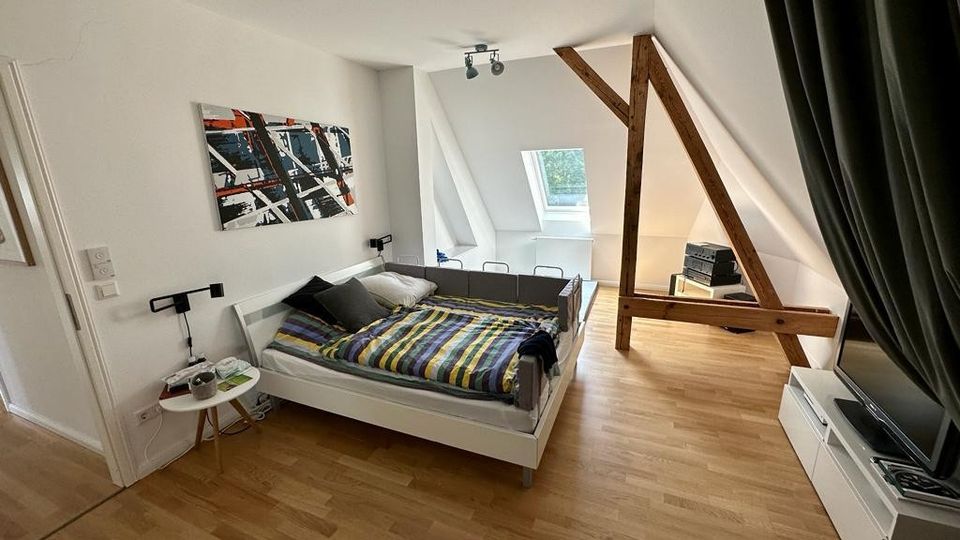 Dachgeschoss-Schlafzimmer mit Holzboden, Schrägdecke und sichtbaren Holzbalken. Das Zimmer enthält ein Bett, ein kleines Nachttischchen, ein TV-Regal sowie ein Dachfenster, das Tageslicht hereinlässt.