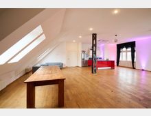 Großzügiger, modern gestalteter Raum mit Dachschrägen und Oberlichtern, einem Holzboden und einer offenen Küche mit roten Schränken. Der Raum enthält einen großen Holztisch, eine graue Couch sowie einen freistehenden Metallträger in der Mitte, und ist von Fenstern mit schwarzen Vorhängen beleuchtet.