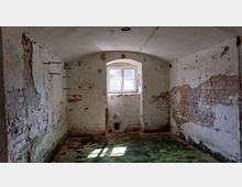 Ein leerer, verfallener Raum mit abblätterndem Putz an den Wänden, teils freiliegendem Ziegelmauerwerk und einem kleinen Fenster, das Licht hereinlässt. Der Boden ist mit feuchten, moosbewachsenen Stellen bedeckt, und die Decke ist gewölbt.