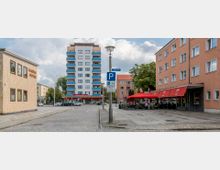 Straßenszene mit mehrstöckigem Wohngebäude im Hintergrund, einem Gebäude mit rotem Sonnendach und Straßencafé auf der rechten Seite sowie einer Apotheke auf der linken Seite. Kopfsteinpflaster, Parkplatzschilder und eine Straßenlaterne prägen den Platz.