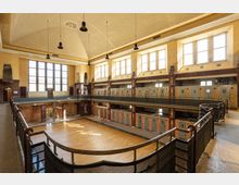 Innenraum eines historischen Schwimmbads mit hohen Decken, großen mehrteiligen Fenstern und gemauerten Wänden. Unten befindet sich eine Parkettfläche anstelle eines Wasserbeckens, umgeben von alten Kabinen mit Türen in hellen Farben, sowie Galerien mit Geländern auf zwei Ebenen.