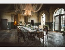 Rustikaler Festsaal in einem alten Industrie- oder Gutshausgebäude mit hohen Decken, abgenutzten Wänden und Holzparkett im Fischgrätmuster. In der Mitte steht ein lang gedeckter Tisch mit Blumenarrangements, darüber hängen ein Kronleuchter und drapierte Stoffbahnen; große Rundbogenfenster und Glastüren lassen Tageslicht herein.