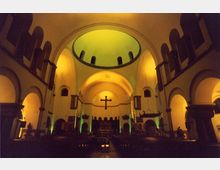 Innenraum einer Kirche mit hohen Rundbögen und einer großen Kuppel. Der Raum ist in warmem gelbem und grünem Licht beleuchtet, und ein großes Kreuz hängt zentral an der Wand über dem Altarbereich.