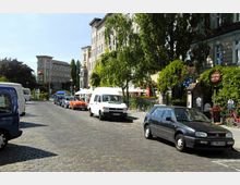 Pflasterstraße in einer städtischen Umgebung mit geparkten Autos, großen Wohngebäuden im Hintergrund und grünen Bäumen entlang des Gehwegs. Rechts ist ein Café mit Außensitzbereich unter Sonnenschirmen zu sehen.