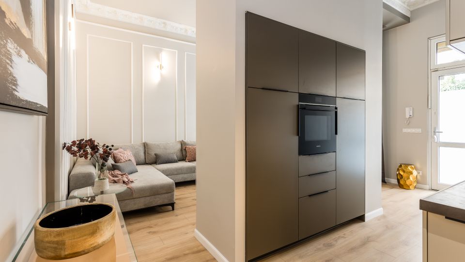 Offener Wohn- und Küchenbereich mit hellem Holzboden, weiß vertäfelten Wänden und Stuckdecke. Links steht ein graues Ecksofa im Wohnbereich, rechts eine moderne dunkle Einbauküchenwand mit integriertem Backofen neben einer Glastür zum Außenbereich.