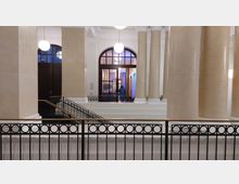 Großes Innenraumfoyer mit hohen, cremefarbenen Säulen und prächtigen Rundbögen. Im Vordergrund ein schwarzes Geländer mit kreisförmigem Muster, im Hintergrund eine Glas-Doppeltür mit Blick auf beleuchtete Wände und eine Person.