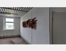 Ein leerer Raum mit weißen Wänden und einer Metallstreifendecke. An einer Wand hängt ein kunstvolles Geflecht aus Naturmaterialien, während ein Fenster mit Blick auf Bäume und Dächer links zu sehen ist.