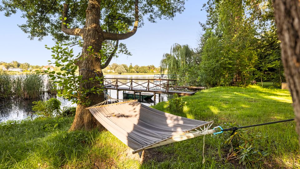 Uferwiese an einem See mit großen Bäumen und einer zwischen zwei Stämmen gespannten Hängematte im Vordergrund. Im Hintergrund führt ein hölzerner Steg mit Geländer ins Wasser, umgeben von Schilf und dichter Ufervegetation.