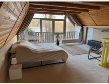 Dachzimmer mit Holzverkleidung und schrägen Wänden, eingerichtet mit einem Bett, einer Stehlampe, einem Teppich und minimalistischem Mobiliar. Große Fenster mit Blick auf einen Balkon und eine natürliche Landschaft mit Bäumen und einem See im Hintergrund.