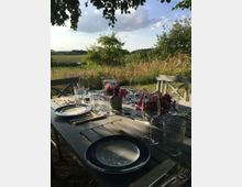 Ein gedeckter Holztisch im Freien mit blauen Tellern, Gläsern, Besteck und Blumen in Vasen, umgeben von einer Wiese und Bäumen unter einem sonnigen Himmel. Im Hintergrund ist eine weitläufige Landschaft mit Feldern und Sträuchern zu sehen.
